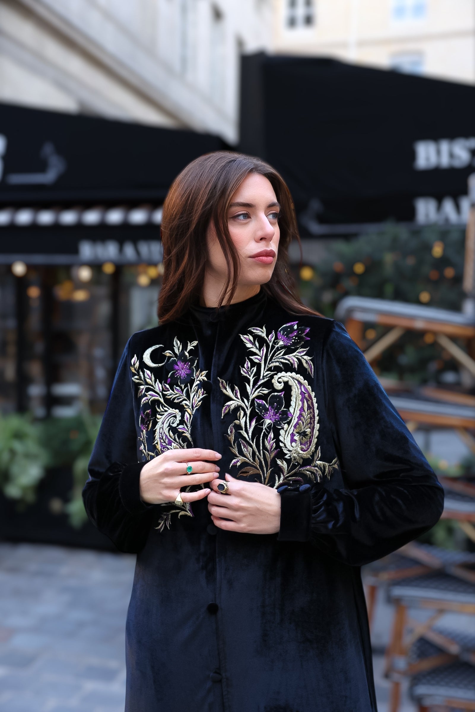 Woman wearing a black velvet dress with intricate gold and purple floral embroidery, photographed in a historic architectural setting in Paris, highlighting artisanal craftsmanship and refined luxury.