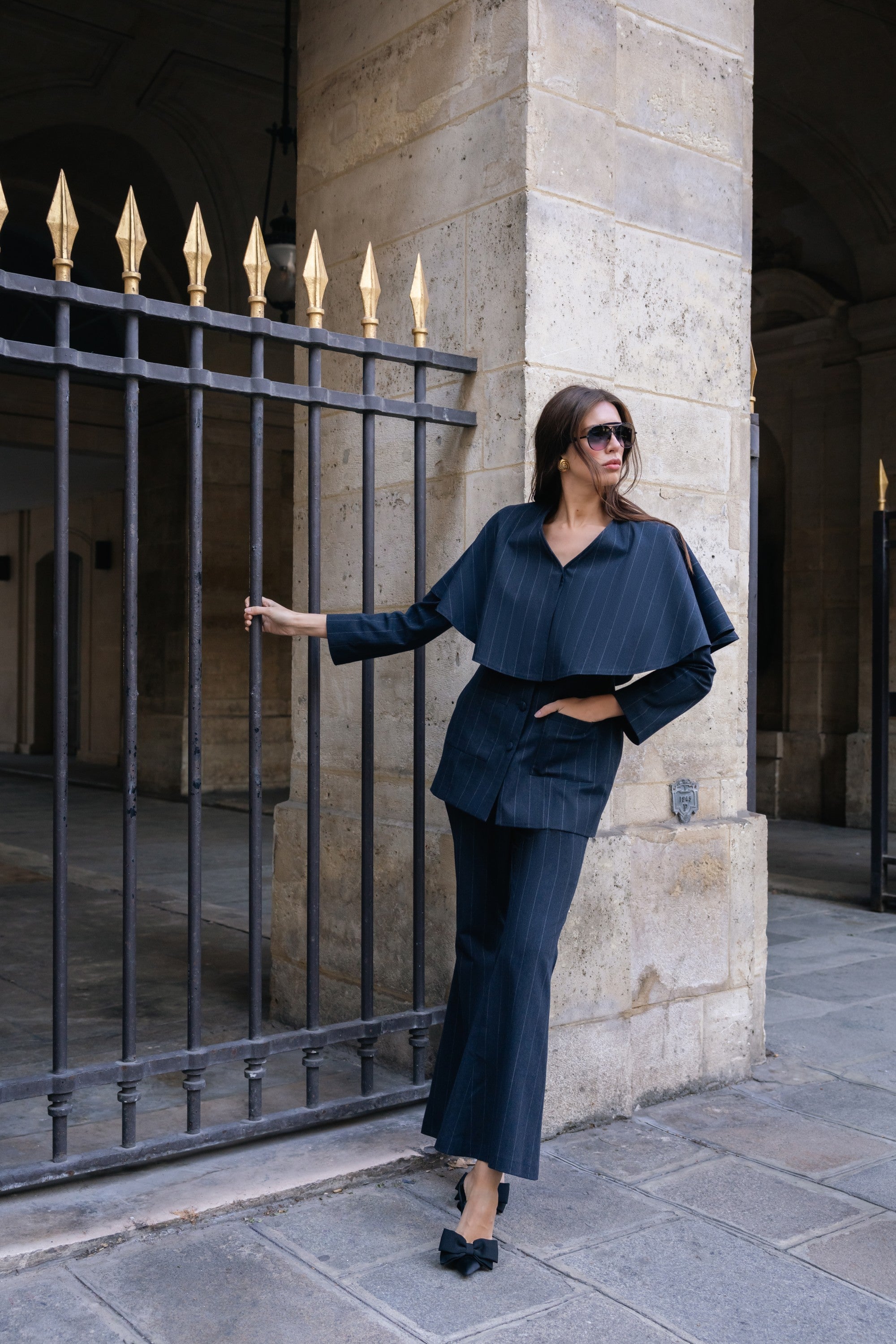 Woman in a pinstripe suit outfit standing next to a stone building with a metal gate in Paris.