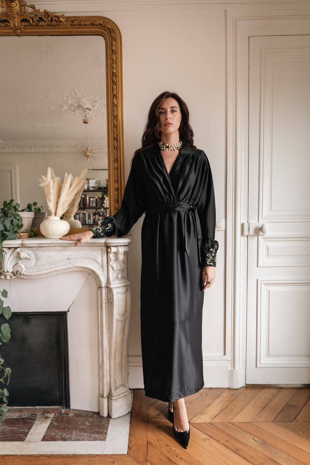 Woman in a black dress standing in a stylish room with a fireplace and decorative mirror.