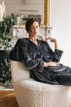 Woman in dark silver velvet suit outfit sitting on a textured chair in a room with decorative elements. Eid outfit, christmas outfit. party outfit