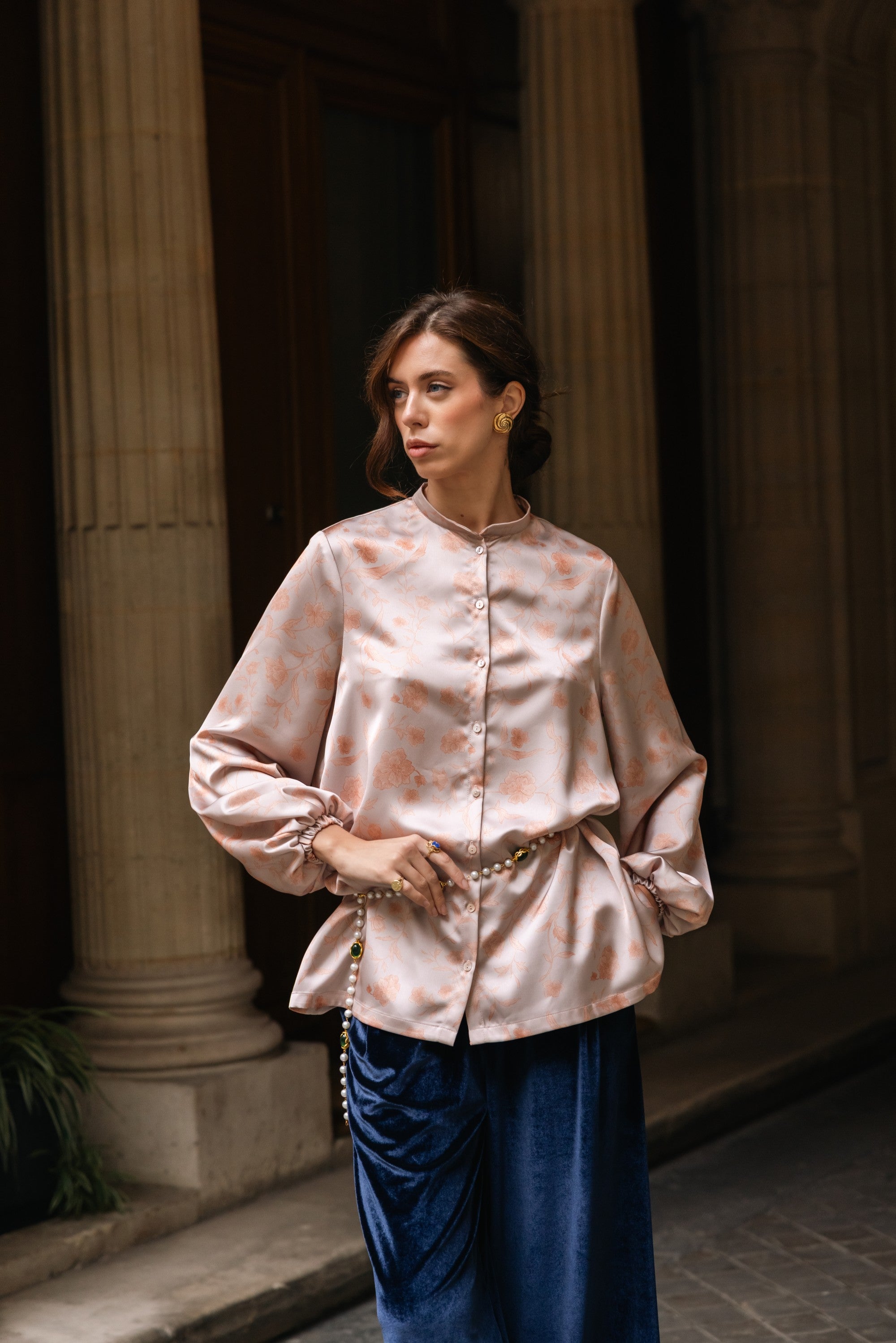 Woman wearing a light pink floral blouse and blue pants standing in front of classical columns.